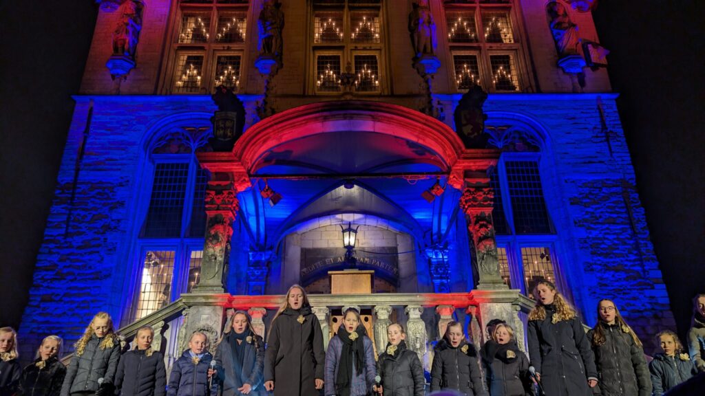 Children’s choir performing in front of Gouda’s historic Town Hall, beautifully illuminated in red and blue, during Gouda Candle Light Festival. Magical Christmas event with candles, music, and festive atmosphere in the heart of Gouda, Netherlands.