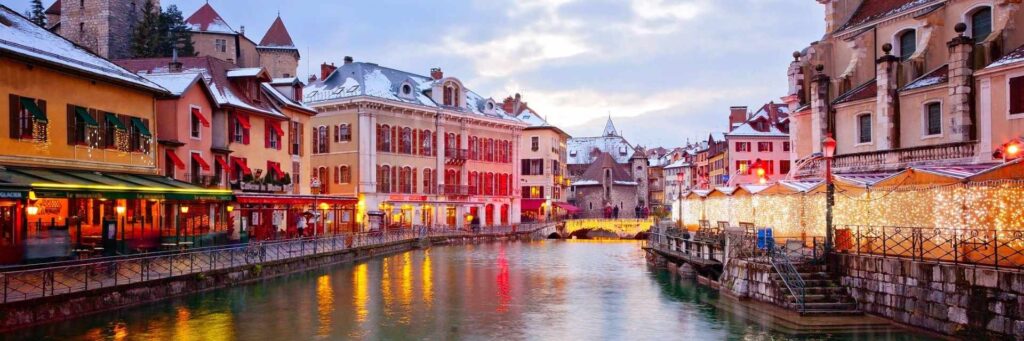Snow-dusted buildings and sparkling Christmas Market stalls lining the canal in Annecy, France. Festive holiday lights reflect on the water, creating a magical winter scene for family travel and seasonal adventures.