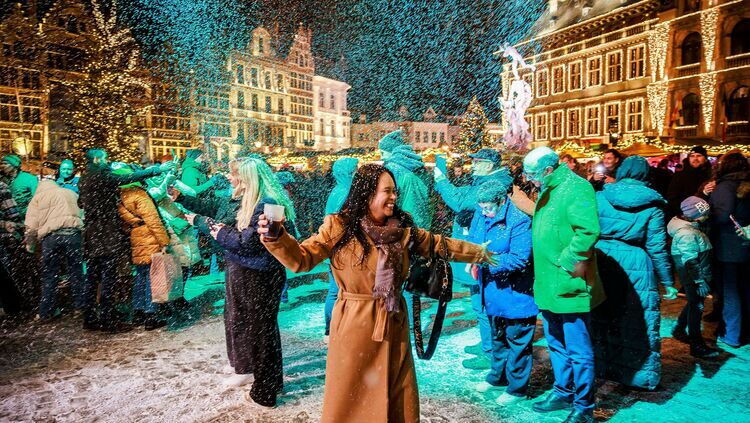 Top 10 Kindvriendelijke Kerstmarkten in Belgiƫ 77 People enjoying artificial snowfall and holding drinks at Antwerp Christmas Market in Belgium, surrounded by illuminated historic buildings and lively holiday crowds on a festive winter night.