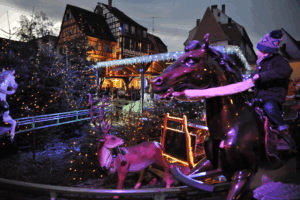 Child riding a carousel horse surrounded by Christmas trees, reindeer decorations, and sparkling holiday lights at Colmar Christmas Market Petite Venise, Alsace, France. A magical winter scene perfect for family travel and festive experiences with children.