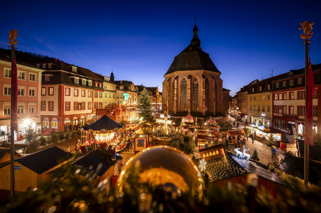 Experience the enchanting Heidelberg Christmas Market with the Church of the Holy Spirit as a glowing backdrop, surrounded by twinkling stalls and festive lights. This magical night scene showcases charming holiday traditions, illuminated decorations, and joyful visitors in Heidelberg’s historic city center