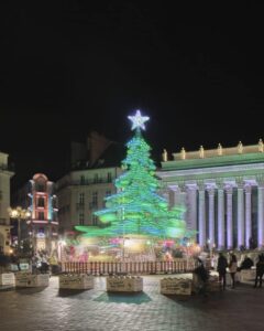 Illuminated green Christmas tree carousel glowing in front of classical buildings at Nantes Christmas Market, with families admiring the festive light installation and enjoying magical winter nights in western France.