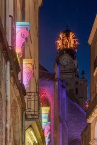 Colorful light installations glowing on historic facades leading toward a beautifully lit church clock tower at night during Nantes Christmas Market, creating a magical festive atmosphere in the old town streets of western France.