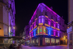Vibrant pink and blue light projections covering a historic corner building above busy café terraces at Nantes Christmas Market, creating a colorful festive atmosphere in the evening streets of western France.