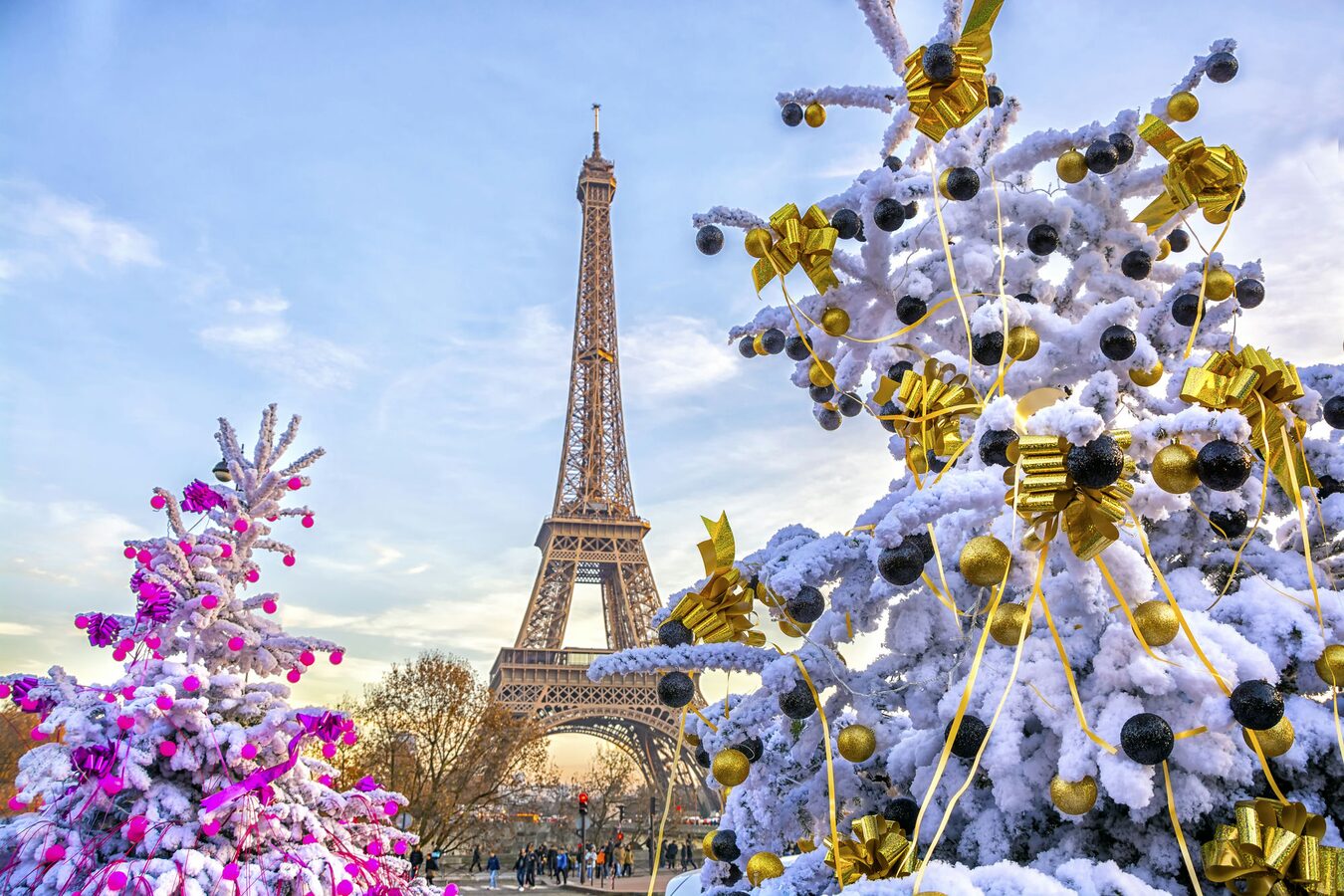 Festive snowy Christmas trees adorned with gold and pink decorations near the Eiffel Tower at Paris Christmas Market, with a clear sky and visitors enjoying the iconic holiday scene. Best for winter travel, family holidays, and magical moments in Paris, France.