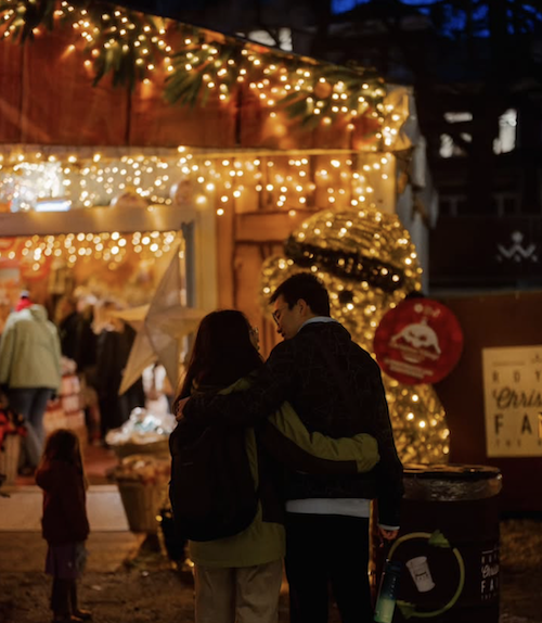 Couple walking arm in arm past a warmly lit Christmas stall at The Hague’s Royal Christmas Fair, with festive lights glowing in the winter evening.
