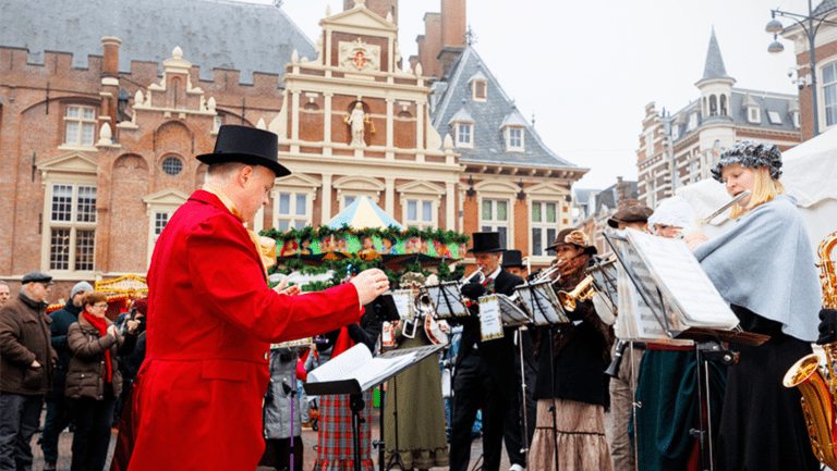 Victorian-costumed musicians perform in front of decorated stalls and historic Dutch architecture at Haarlem Christmas Market. Guests enjoy live holiday music, festive atmosphere, and traditional market celebrations in the heart of Haarlem, Netherlands.
