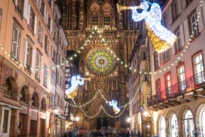 Strasbourg’s festive Christmas Market street lit up with sparkling angel decorations and vibrant lights, leading to the majestic Notre-Dame Cathedral rose window in the evening. A magical holiday destination for families and winter travelers in Alsace, France.
