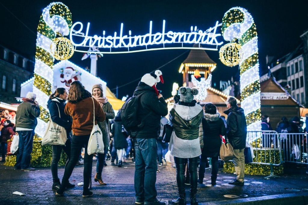 Visitors entering the illuminated Christkindelsmärik gate at Strasbourg Christmas Market, surrounded by festive lights and holiday decorations on a vibrant winter night. Perfect for family holidays and joyful celebrations in Alsace, France.