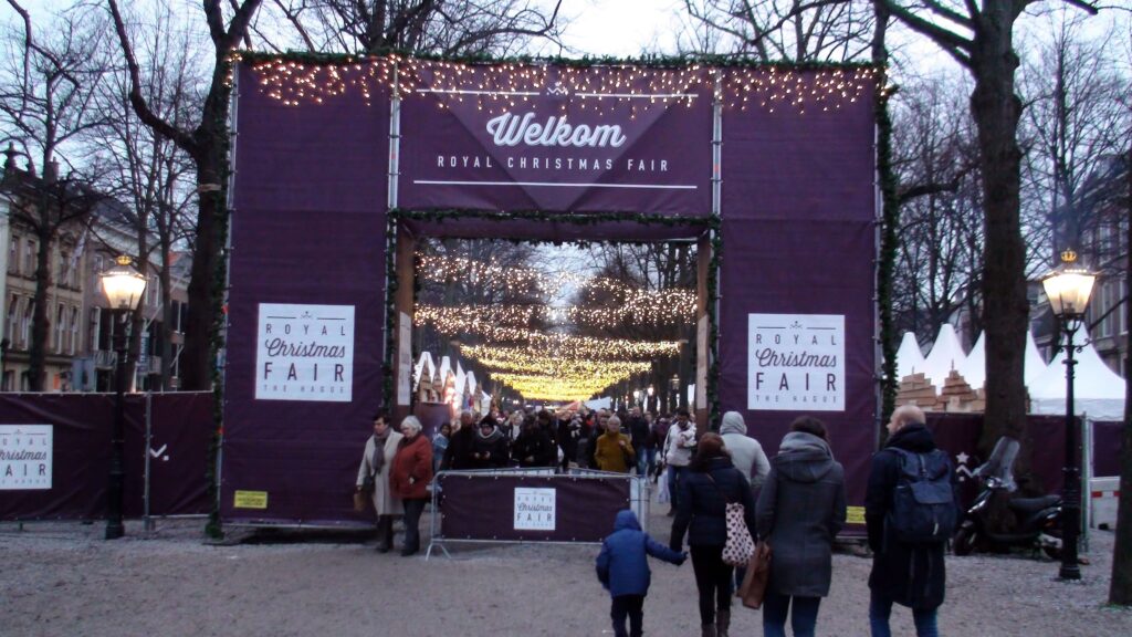 Families and holiday visitors enter The Hague Royal Christmas Fair beneath sparkling canopy lights and welcoming signs. Festive entrance to the Christmas market lined with decorated trees and cheerful winter atmosphere in central The Hague, Netherlands.