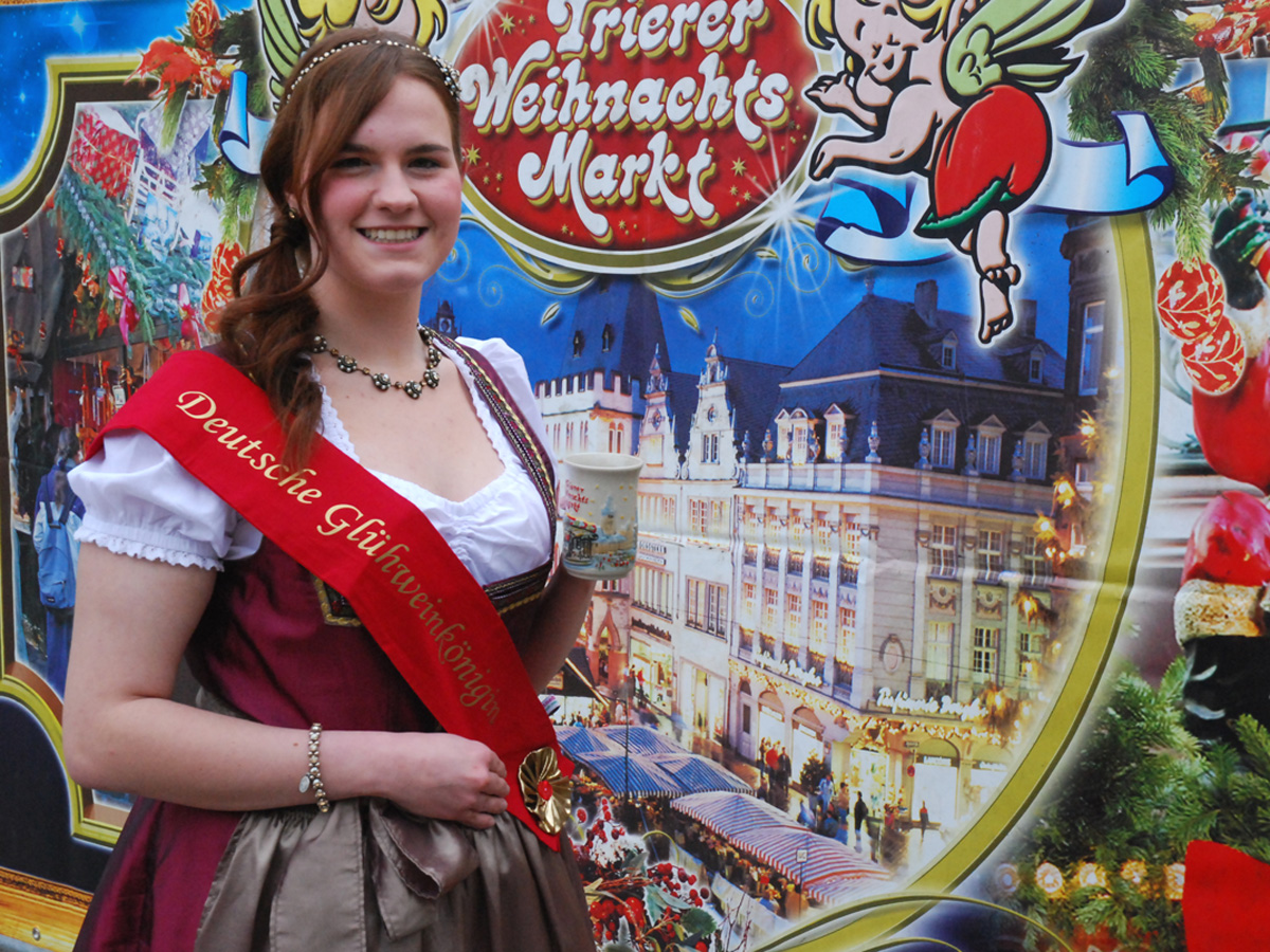 Celebrate tradition at Trier Christmas Market with the German Mulled Wine Queen featured in front of a colorful holiday backdrop. This cheerful scene highlights festive costumes, Glühwein culture, and the unique charm of one of Germany’s oldest Christmas markets.
