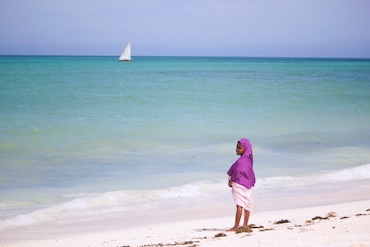 Vrouw in paarse doek wandelt over het witte strand van Zanzibar met een zeilboot op de turquoise zee – rust en lokale cultuur aan de Indische Oceaan.